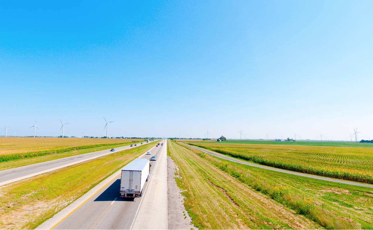 A truck driving down a road with green fields on either side and wind turbines in the background