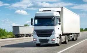 A truck with box van trailer drives down the road, with another trailer in the background.